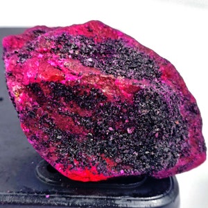 Könnte beinhalten: Ein großer, rauer, rosafarbener Rubin-Edelstein mit schwarzen Einschlüssen. Der Rubin liegt auf einer schwarzen Oberfläche.