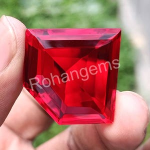 Puede incluir: Una piedra preciosa roja oscura facetada con forma cuadrada. La piedra preciosa está tallada en un estilo único con una parte superior puntiaguda y una base plana. La piedra preciosa se sostiene en una mano, y la palabra "Rohangems" es visible en el fondo.
