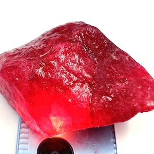 Op de afbeelding: Een ruwe, ongeslepen robijn edelsteen met een dieprode tint. Het oppervlak van de steen heeft een textuur, met variaties in kleur en lichtreflectie. De edelsteen wordt gepresenteerd tegen een witte achtergrond.