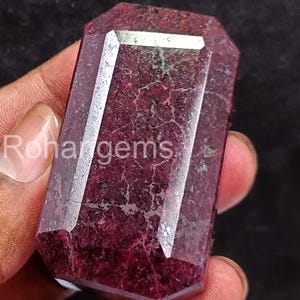 May include: A faceted, rectangular gemstone with a deep red hue. The gem has a polished surface with visible internal patterns and the text "Rohargems" on the side. The gem is held against a dark background.