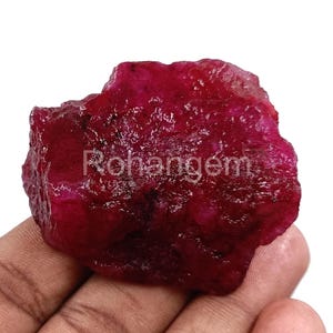 Puede incluir: Una gema de rubí en bruto, sin cortar, de color rojo intenso. La superficie tiene un aspecto texturizado. El texto "Rohangem" es visible en la superficie de la gema.