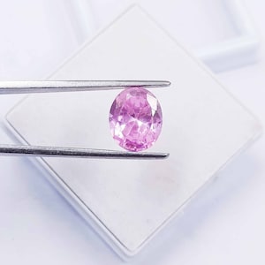 Può includere: Una pietra preziosa ovale di colore rosa pallido, tenuta da una pinzetta. La pietra preziosa è sfaccettata.