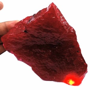 May include: A large, rough, red gemstone with a translucent, glassy appearance. The gemstone is irregularly shaped and has a slightly pointed top.