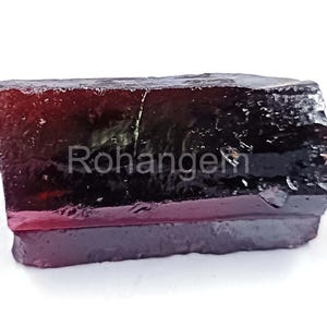 Op de afbeelding: Een ruwe, rechthoekige edelsteen met een diepe bordeauxrode en zwarte kleurovergang. De steen heeft een ruwe, natuurlijke textuur en de tekst "Rohangem" is zichtbaar op het oppervlak. Dit mineraal specimen is waarschijnlijk een toermalijn of een vergelijkbare kristal.