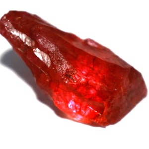 May include: A raw, uncut gemstone with a deep red hue. The gem has a rough, textured surface and irregular shape, catching the light to create highlights and shadows. The stone is set against a white background, emphasizing its color and form.