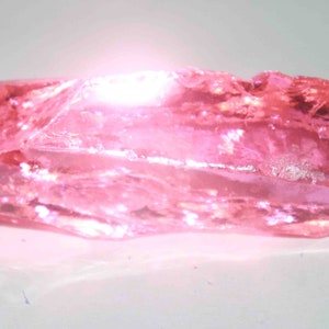 May include: A translucent, rectangular pink gemstone with a rough, uneven surface. The stone has a crystalline structure and reflects light, creating a shimmering effect. The overall color is a vibrant, rosy pink.