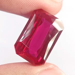 May include: A vibrant, deep red gemstone with a rectangular emerald cut. The stone features multiple facets that reflect light, creating a brilliant, translucent appearance with sharp edges and a polished, smooth surface throughout the entire piece.