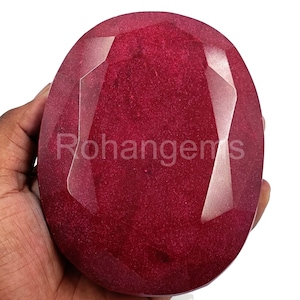 Puede incluir: Una gran gema roja oscura con un corte facetado. La gema tiene una forma ligeramente ovalada y una superficie brillante y pulida.
