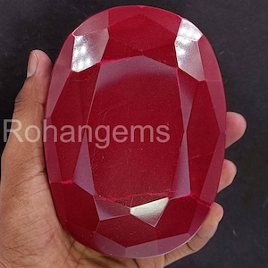 May include: A large, faceted, oval-shaped ruby gemstone. The deep red gem is held in a hand, showcasing its polished surfaces and intricate cuts. The text "Rohangems" is visible on the gem's surface.
