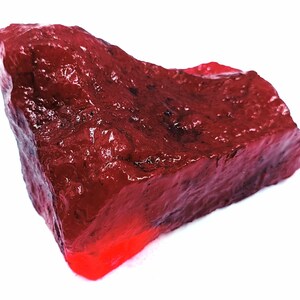 May include: A rough-cut, deep red gemstone with a slightly glossy surface. The stone has a natural, irregular shape and a vibrant, saturated color. The gemstone is set against a clean white background.