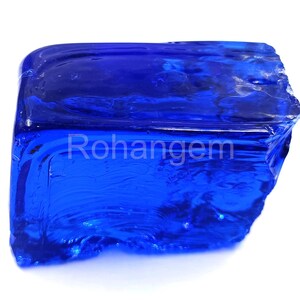 May include: A vibrant, translucent, deep blue gemstone with a rough, irregular shape. The surface reflects light, showcasing the gem's internal structure. The text "Rohangem" is visible on the surface. The gem is set against a white background.