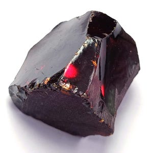 May include: A dark burgundy gemstone with an irregular shape. The surface is glossy and reflective, with flashes of red and gold. The stone appears to be uncut, displaying natural textures and a raw, unpolished appearance.