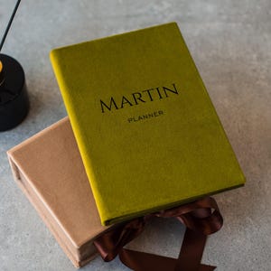 May include: An olive green planner with the name "MARTIN" and the word "PLANNER" printed in black. A brown box and a black diffuser with reeds are also in the image. A brown ribbon is tied around the box.
