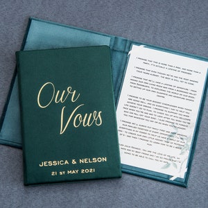 Puede incluir: Un libro de votos de terciopelo verde con letras doradas que dicen "Our Vows" y "Jessica & Nelson 21st May 2021". El libro está abierto para revelar una página blanca con votos escritos en tinta negra.