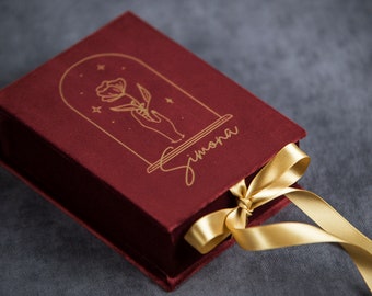 Könnte beinhalten: Eine burgunderfarbene Samtbox mit einem goldenen Band. Die Box hat ein goldenes Design mit einer Blume in einer Kuppel mit Sternen und dem Namen "Simona" darunter.