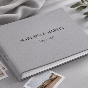 May include: A light gray guest book with a fabric-like texture, featuring the black printed text "MARLENE & MARTIN" and "July 7, 2027" on the front cover, with the words "GUEST BOOK" printed in black on the spine of the book.