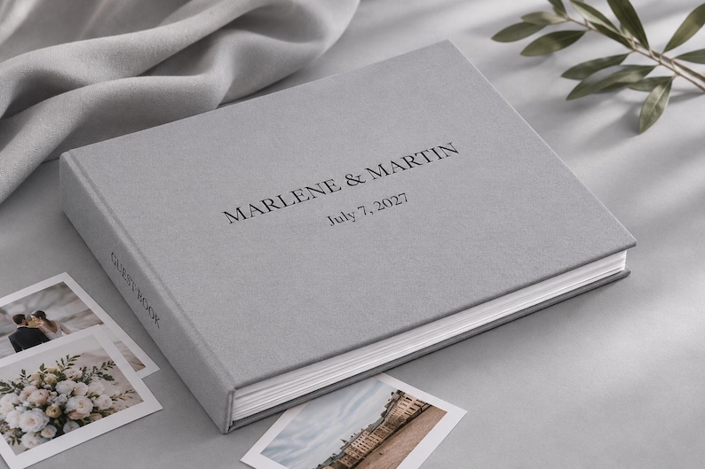 May include: A light gray guest book with a fabric-like texture, featuring the black printed text "MARLENE & MARTIN" and "July 7, 2027" on the front cover, with the words "GUEST BOOK" printed in black on the spine of the book.