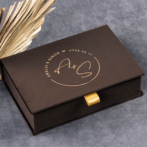 May include: A brown box with a gold ribbon and a gold inscription that reads "Amelia & Simon 2023.03.15" with the initials "A+S" in a circle.