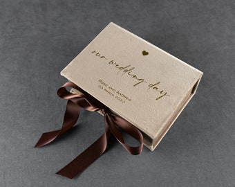 Könnte beinhalten: Eine beige Samtbox mit einem braunen Band darum gebunden. Die Box trägt den Text "our wedding day" und "ROSE AND ANDREW 03 MARCH 2023" in goldenen Lettern.
