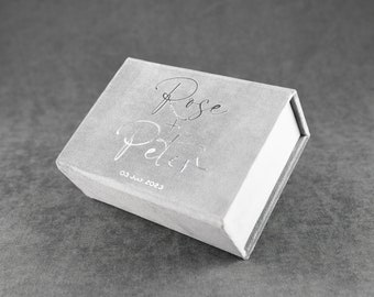 May include: A grey velvet box with the words "Rose + Peter" and the date "03 Jul 2023" embossed in silver.