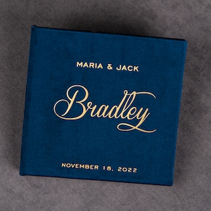 Puede incluir: Una caja de terciopelo azul marino con letras doradas. La caja dice "Maria & Jack" y "Bradley" en una fuente cursiva. La fecha "18 de noviembre de 2022" está impresa debajo del nombre.