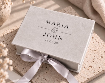 Könnte beinhalten: Eine rechteckige Geschenkbox aus hellgrauem Samt mit Klappdeckel und Satinband. Auf dem Deckel ist der Text MARIA & JOHN sowie das Datum 16 07 28 in einer dunklen Serifenschrift aufgedruckt. Die Box hat eine weiche, strukturierte Oberfläche.