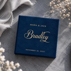 May include: A square navy blue velvet box featuring gold foil text. The top of the box is printed with the names MARIA & JACK, the name Bradley in a large script font, and the date NOVEMBER 18, 2027, centered on the surface.