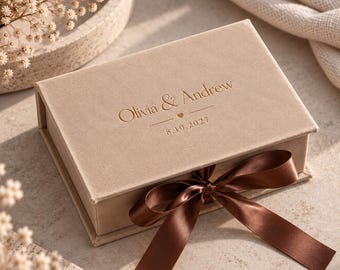 May include: A rectangular beige velvet gift box featuring a hinged lid with the embossed text Olivia & Andrew, a small heart icon, and the date 8.10.2027. The box is secured with a dark brown satin ribbon tied in a bow at the front centre.