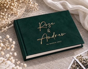 May include: A dark green velvet-covered photo album with gold foil lettering on the front cover that reads Rose & Andrew and the date 03 MARCH 2027. The album has a rectangular shape with a thick spine and white pages visible along the edge.