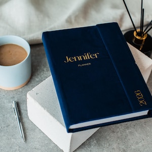 Op de afbeelding: Een marineblauwe planner met de naam "Jennifer" in gouden letters en het jaar "2026" op de rug. Een lichtblauwe mok met koffie en een zilveren pen zijn ook aanwezig. De planner ligt op een wit blok.