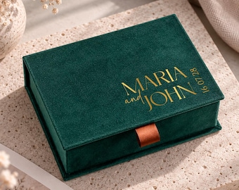 May include: A rectangular dark green velvet jewellery box featuring gold foil lettering that reads MARIA and JOHN with the date 16 07 28. The box has a small brown ribbon pull tab on the front edge and a hinged lid design with a soft texture.