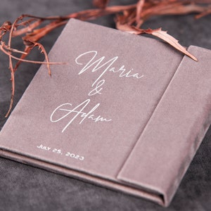 May include: A light pink velvet box with the text "Maria & Adam" and the date "July 25, 2023" printed in white.