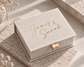 Könnte beinhalten: Eine rechteckige Schmuckschatulle aus beigem Samt mit einer goldfarbenen Zuglasche an der vorderen Schublade. Auf der Oberseite der Schatulle sind die Namen James & Sarah in einer goldfarbenen Schreibschrift mittig aufgedruckt.