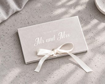 May include: A rectangular, light beige velvet-textured box featuring the white printed text "TO THE NEW Mr and Mrs" on the cover. A white satin ribbon is tied in a bow around the center of the box, securing the lid with a small metal fastener.