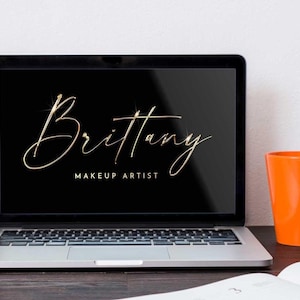 May include: A silver laptop computer with a black screen displaying a gold script logo that reads "Brittany Makeup Artist". The laptop is on a wooden table next to an orange mug.