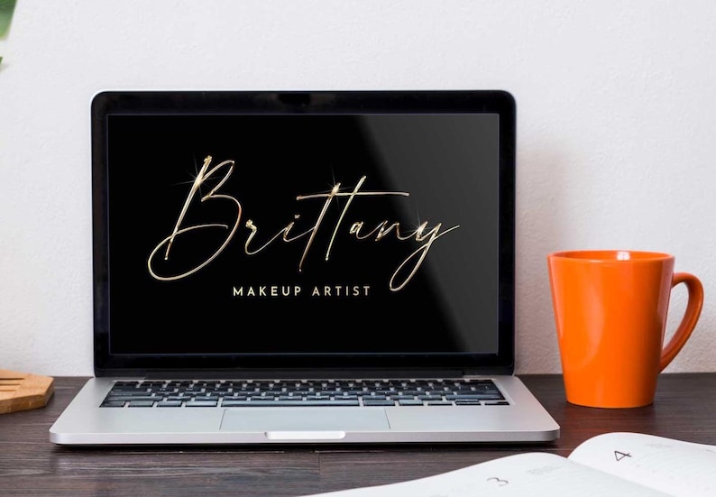 May include: A silver laptop computer with a black screen displaying a gold script logo that reads "Brittany Makeup Artist". The laptop is on a wooden table next to an orange mug.
