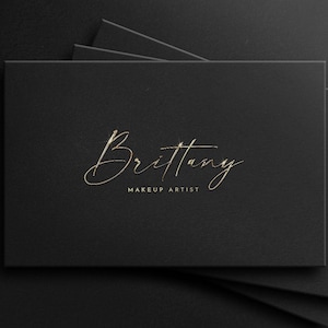 May include: A black business card with gold lettering that reads "Brittany Makeup Artist".