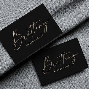 May include: Two black business cards with gold lettering. The text on the cards reads "Brittany Makeup Artist".