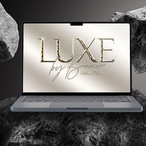 May include: A laptop computer displays the text "LUXE by Denise HAIRSTYLIST" on its screen. The word "LUXE" features a leopard print pattern. The laptop is gray and rests on a dark, textured surface, with large rocks in the background.