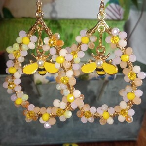 Puede incluir: Aretes de aro dorados con un dije de abeja amarilla y cuentas rosadas y amarillas. Los aretes están decorados con pequeñas flores blancas.