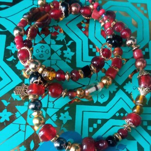 Peut inclure: Un bracelet multi-rangs avec des perles rouges, noires et dorées. Le bracelet comprend un charme en forme de main avec un œil dans la paume.