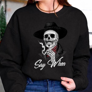 Puede incluir: Sudadera negra con un gráfico detallado de una calavera con sombrero y fumando un cigarrillo. La calavera tiene un pañuelo rojo y las palabras "Say When" están escritas en blanco debajo.