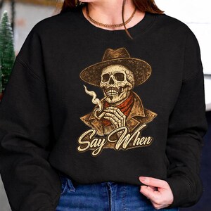 May include: Black sweatshirt featuring a skull wearing a cowboy hat, a red bandana, and a brown jacket, smoking a cigarette. The phrase "Say When" is written in a stylized font below the skull.