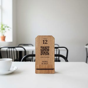 Puede incluir: Un letrero de mesa vertical de madera marrón claro con una base marrón oscuro. El letrero presenta el texto "TABLE 12", un código QR cuadrado, el texto "SCAN TO ORDER" y el nombre de la marca "NORD & SALT" impresos en negro.