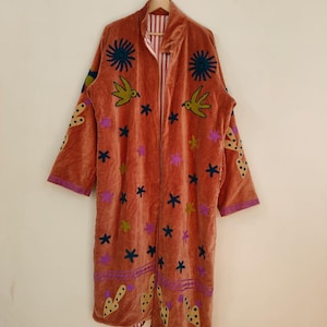 May include: A long, rust-colored velvet robe with embroidered details. The robe features a floral and bird design in shades of green, purple, and yellow. The robe is hanging on a wooden hanger against a neutral background.