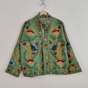May include: A green embroidered jacket with a floral design. The jacket features red, blue, and yellow flowers and leaves. The jacket is hanging on a wooden hanger against a white background.