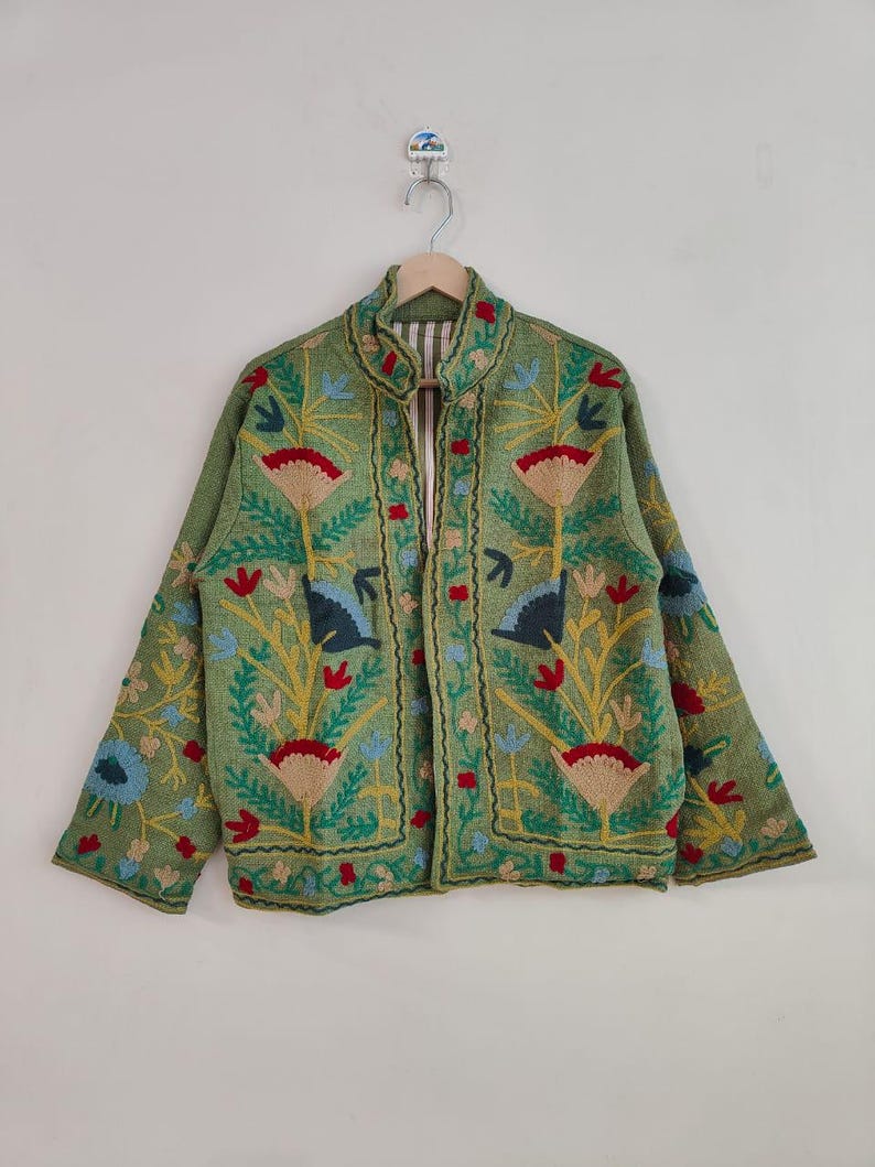 May include: A green embroidered jacket with a floral design. The jacket features red, blue, and yellow flowers and leaves. The jacket is hanging on a wooden hanger against a white background.