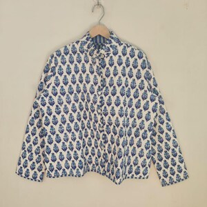May include: A white shirt with a blue floral design, hanging on a wooden hanger. The long-sleeved shirt has a collared neckline and button closures. The floral pattern is repeated across the fabric. The background is a neutral colour.