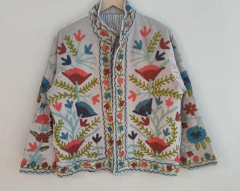 May include: A light-colored jacket with colorful floral embroidery. The jacket features red, blue, green, and orange flowers and leaves. The jacket is hanging on a wooden hanger.