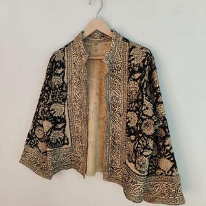 K&ouml;nnte beinhalten: Eine schwarz-beige gemusterte Jacke mit floralem Design h&auml;ngt an einem Holzb&uuml;gel. Die Jacke hat einen hohen Kragen und ist vorne offen, wodurch das beige Innere sichtbar wird. Das florale Muster wiederholt sich auf der gesamten Jacke.